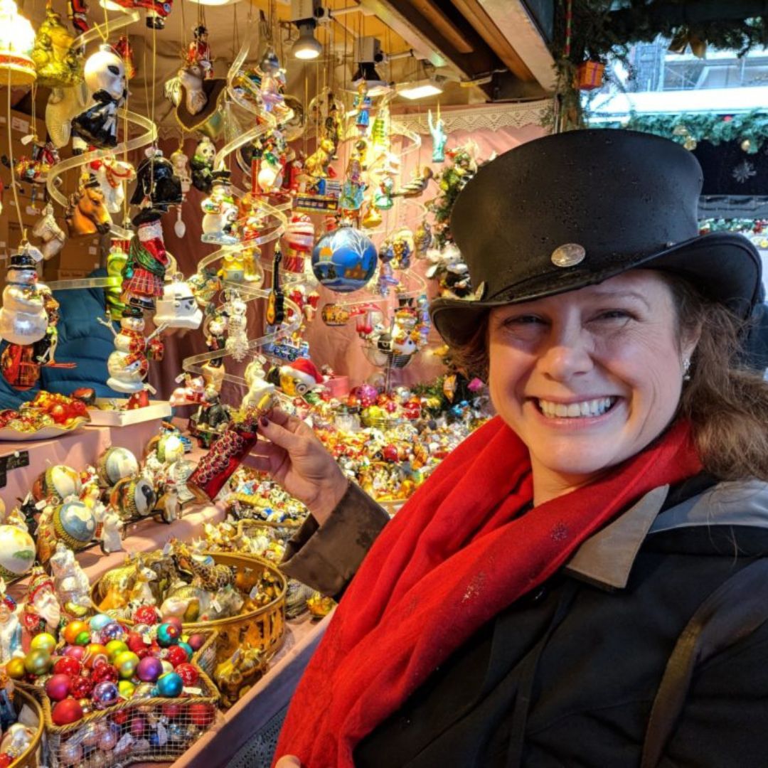 Woman smiling in a black hat and red scarf holding a decorative ornament in front of a vibrant holiday market display filled with colorful ornaments and festive decorations.