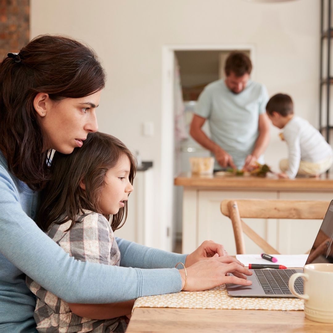 Mother and daughter working on a laptop at home while father prepares food in the background, illustrating remote work and family life during the pandemic.