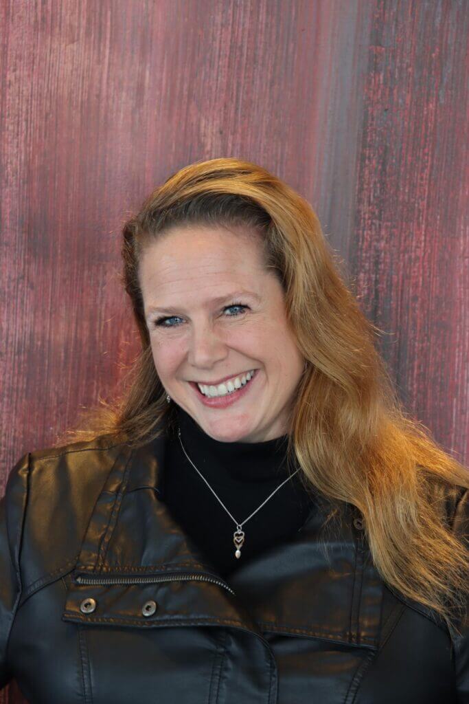 Kristen David smiling in a black leather jacket against a textured red background, representing Upleveling Your Business workshops.