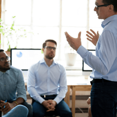 Business meeting with a speaker gesturing, engaged audience, and computer screens displaying charts in a modern office setting.
