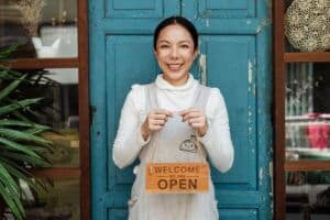 Smiling woman holding an "Open" sign in front of a blue door, representing small business empowerment and welcoming customers.