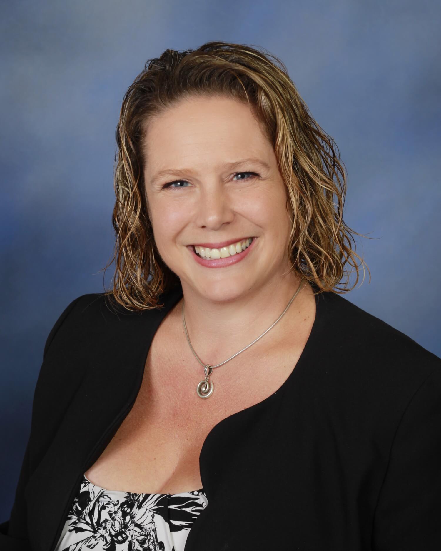 Kristen David smiling in a professional headshot, wearing a black blazer and a floral top, against a blue background, representing her role as a mentor and business coach.