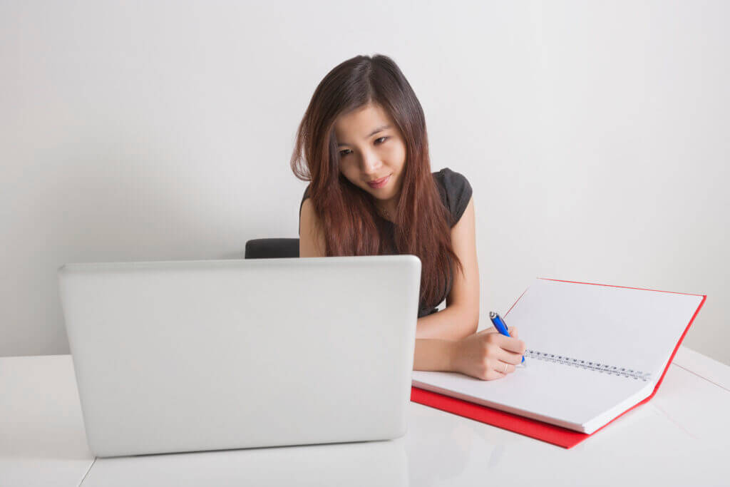 Young woman writing in a notebook while looking at a laptop, representing business coaching and strategic planning for growth.