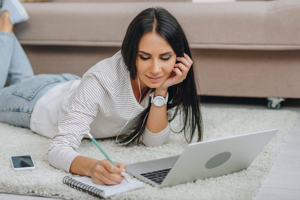 Woman lying on a rug, using a laptop and taking notes in a notebook, illustrating effective business management and learning strategies.