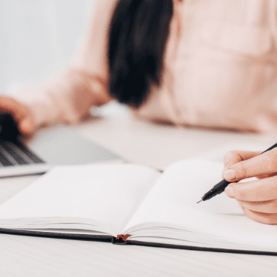 Person writing in a notebook with a pen while using a laptop, representing content creation and marketing strategy development.