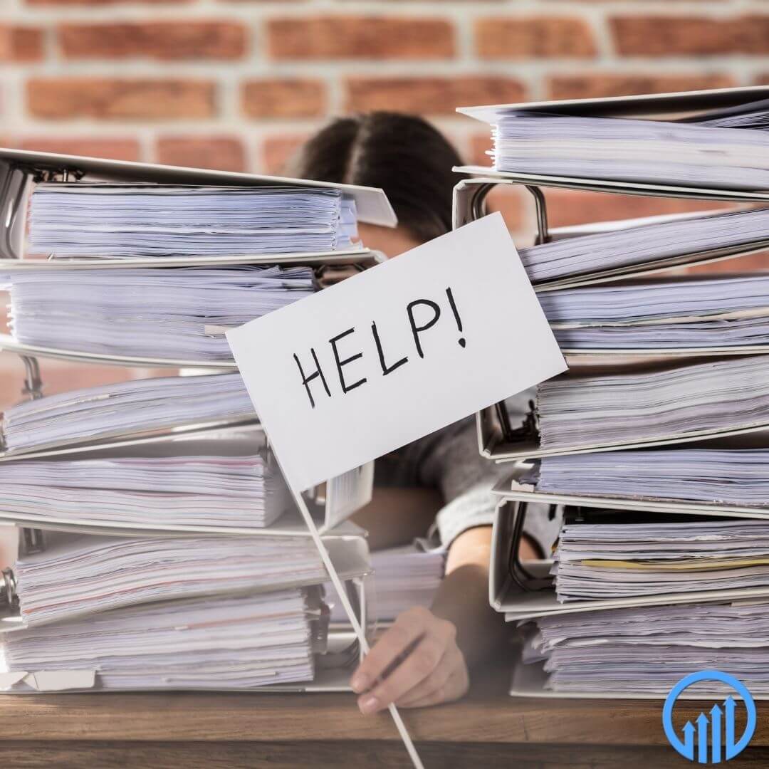 office worker stuck behind two stacks of binders filled with work holding a sign that says "help!"