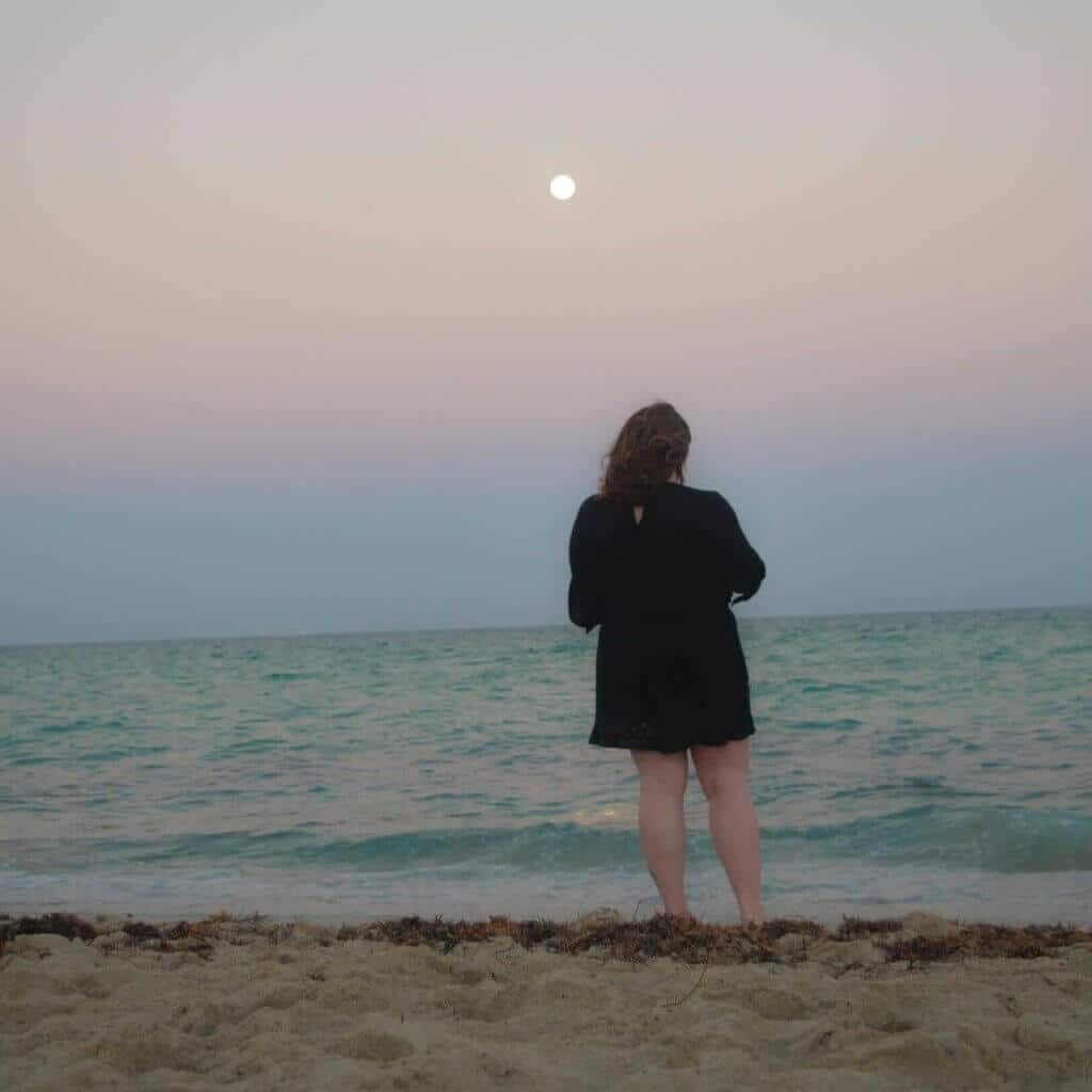 Woman standing on a beach at sunset, gazing at the ocean and moon, reflecting tranquility and connection to nature.