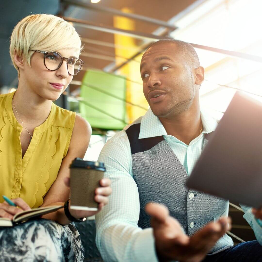 Business professionals discussing ideas in a modern workspace, with one holding a coffee cup and the other a tablet, emphasizing collaboration and planning for new opportunities.