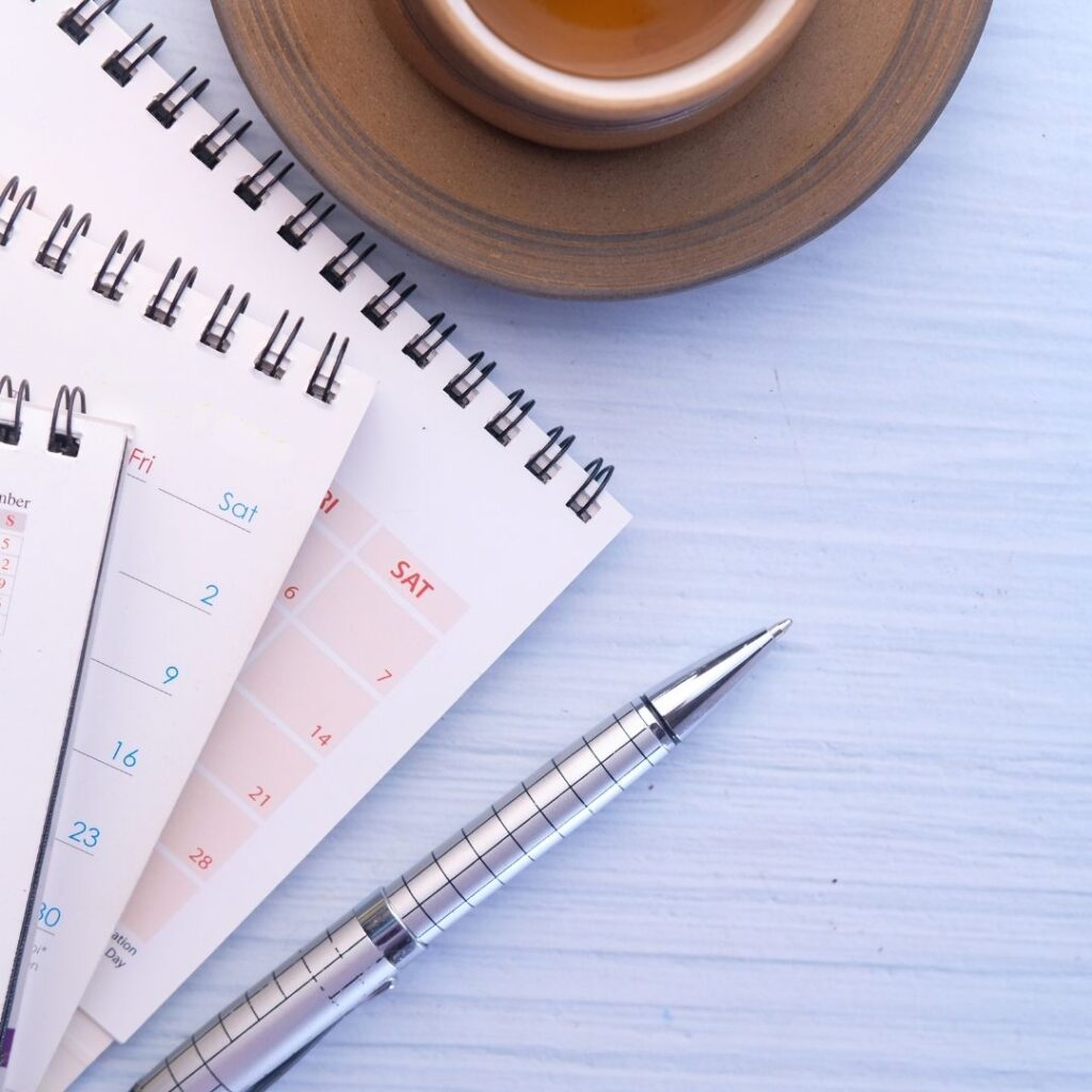 Calendars and notebooks stacked on a light blue surface with a silver pen and a cup of tea, symbolizing effective business planning and organization for year-end sales.