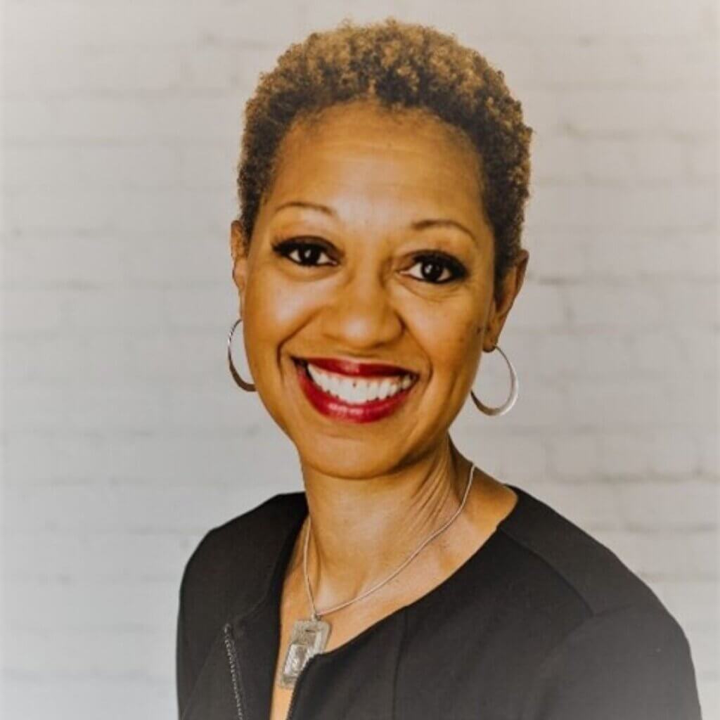 Michele Fuller, smiling sales coach, wearing a black top and necklace, with short hair and hoop earrings, against a light brick background.