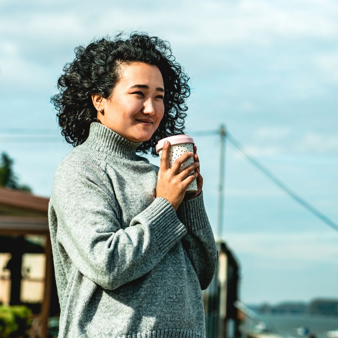 Woman in a cozy sweater holding a coffee cup, smiling outdoors, reflecting the joys and challenges of small business ownership.
