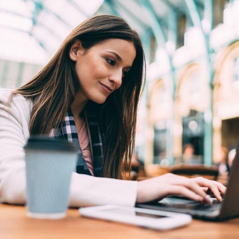 A smiling professional woman working at her laptop with her phone and coffee nearby