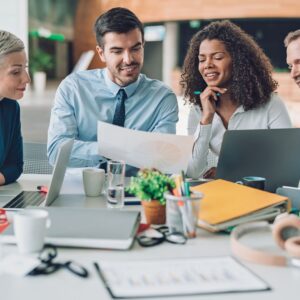 Group of diverse professionals collaborating around a table, discussing marketing strategies and reviewing documents, with laptops and stationery visible, reflecting teamwork in business growth.