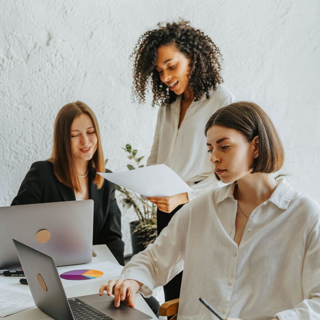 Three women collaborating in a modern office setting, analyzing data on laptops and discussing marketing strategies, emphasizing teamwork and effective communication for marketing plans.