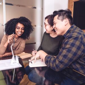 Three diverse business professionals collaborating at a table, discussing strategies and ideas, with a tablet and notebooks visible, reflecting teamwork and support for entrepreneurs.