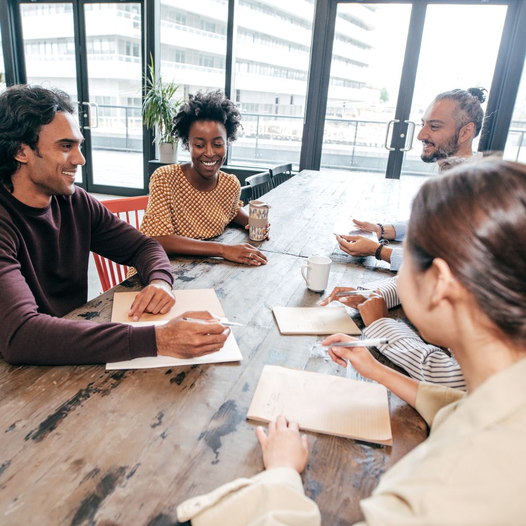 Group of diverse individuals collaborating at a table, discussing ideas and taking notes, highlighting teamwork and communication in a business setting.