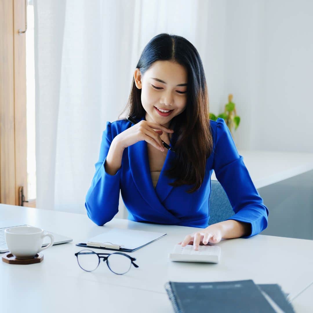 Woman in a blue blazer smiling while working at a desk with a calculator, notepad, and glasses, representing solopreneurship and business growth strategies.