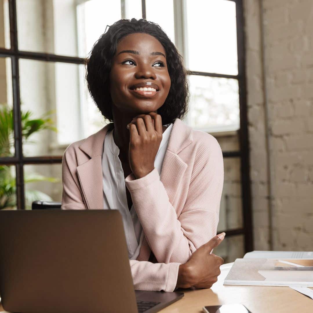 Smiling woman in a pink blazer contemplating at a desk with a laptop, symbolizing strategic growth and collaboration for small business teams.