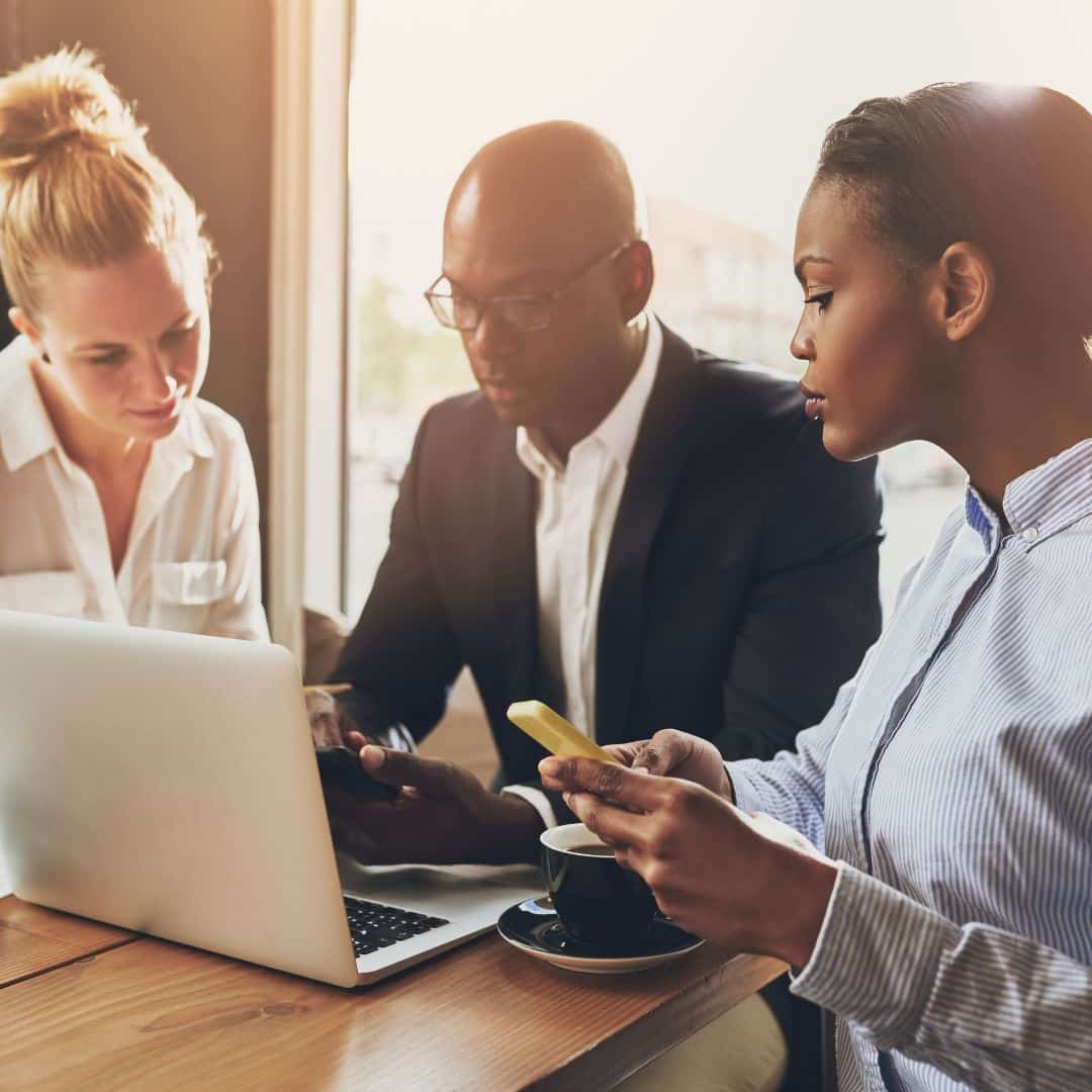 Three professionals collaborating over a laptop in a coffee shop, discussing business strategies and team building, with a cup of coffee on the table.