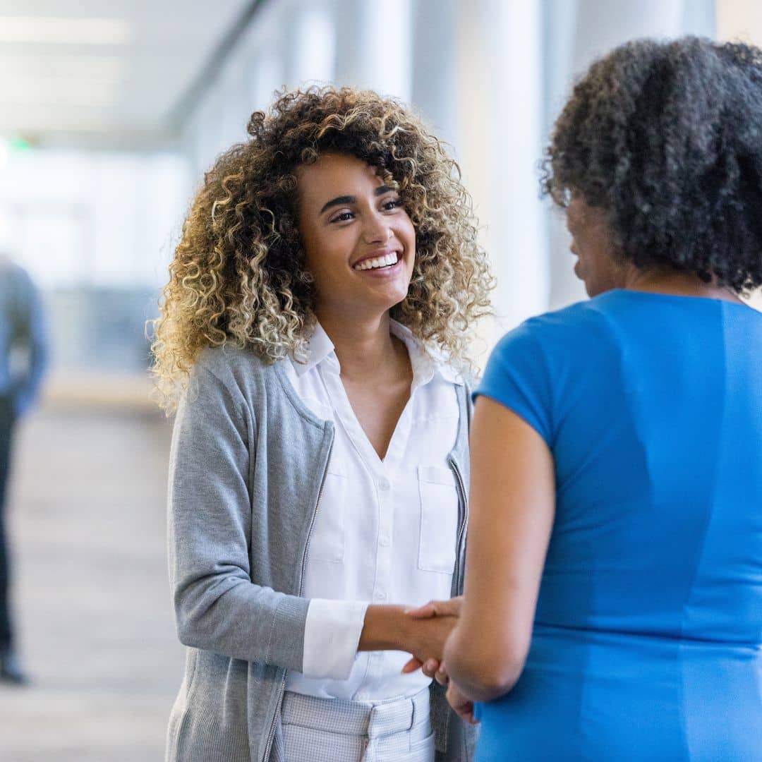 Smiling woman in a gray cardigan shaking hands with another woman in a blue dress, symbolizing teamwork and successful onboarding in a professional setting.