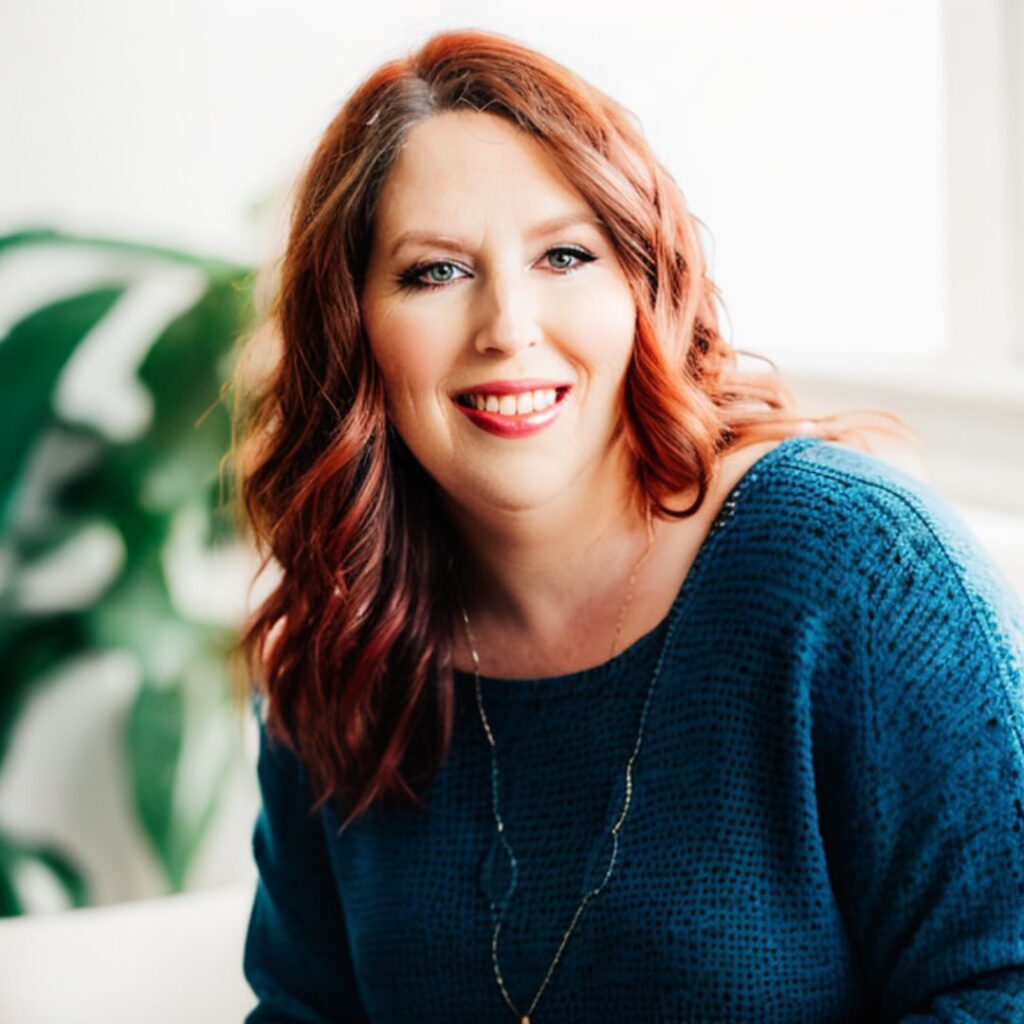 Jen Duncan, marketing and copy coach, smiling in a blue sweater with red hair, surrounded by greenery, emphasizing her expertise in marketing and content strategy.