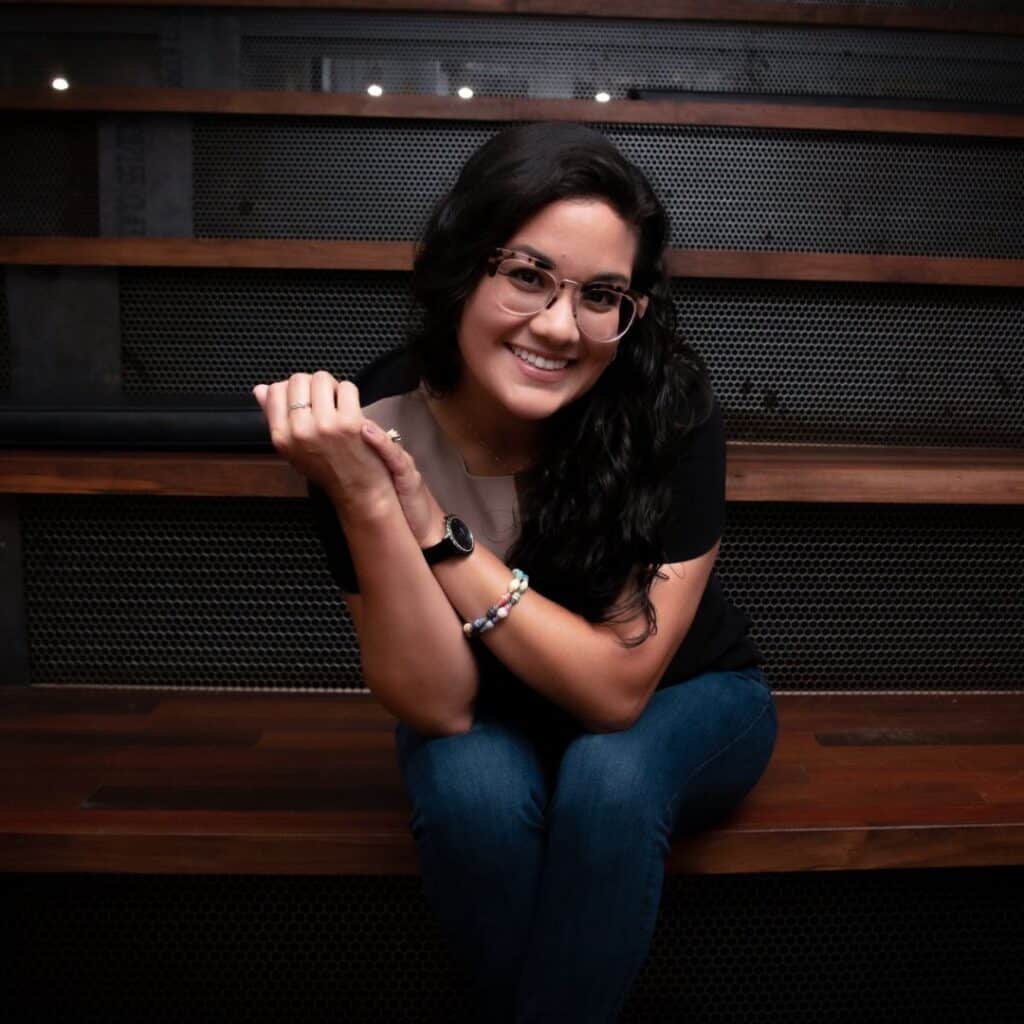 Coach Andrea Rotondo smiling while sitting on wooden stairs, wearing glasses and a black shirt, showcasing her approachable personality as a financial controls coach.
