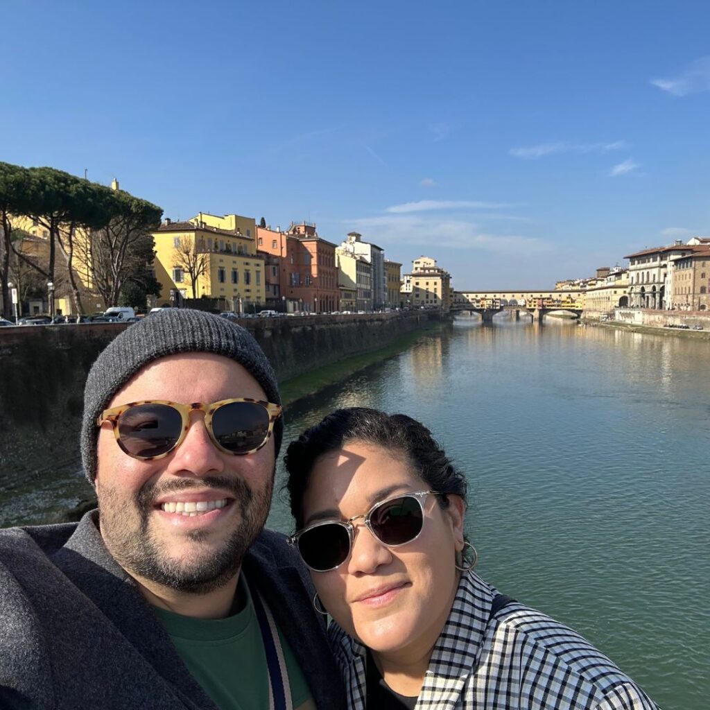 Couple smiling in sunglasses on a bridge with the Arno River and colorful buildings in Florence, Italy.