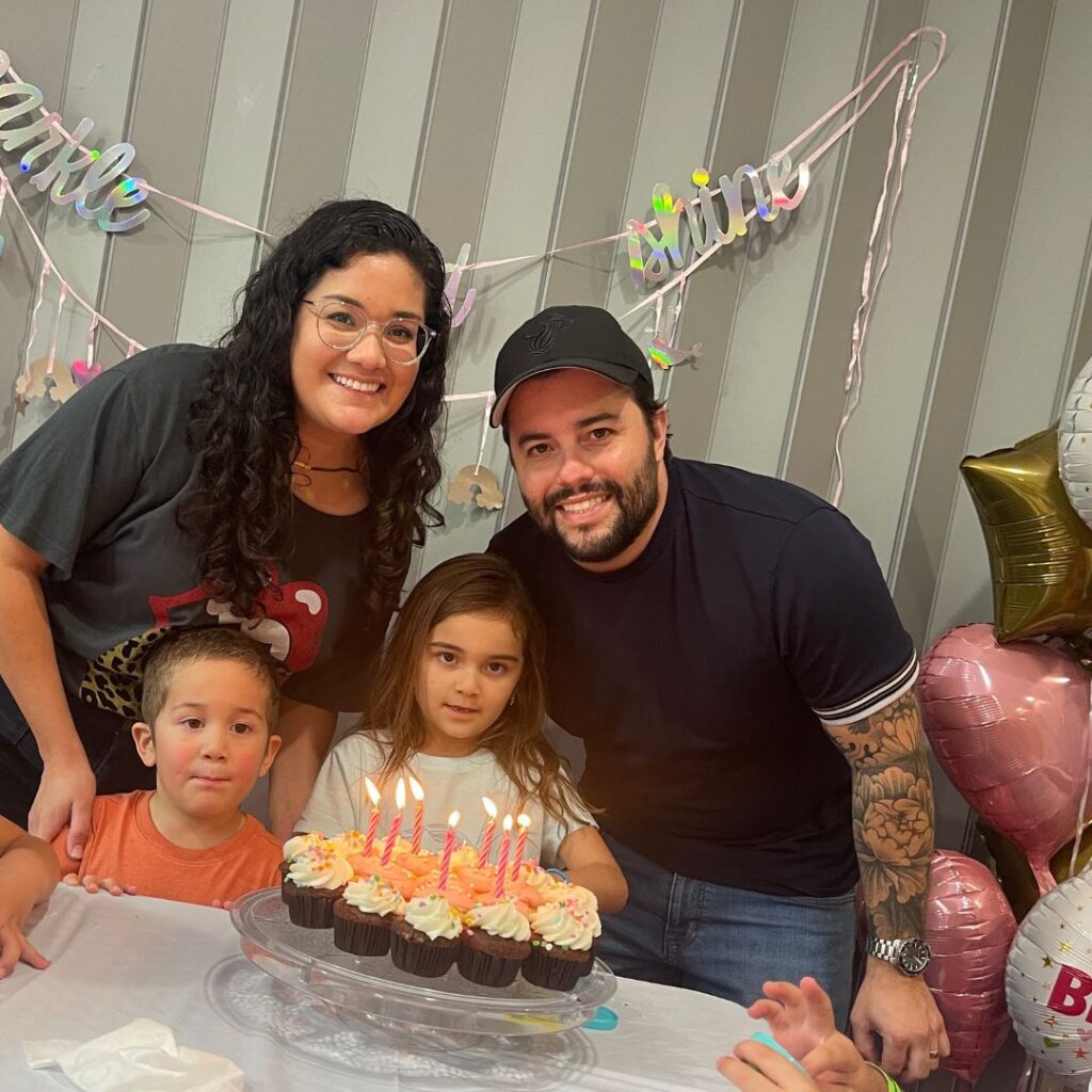 Family celebration with a woman and a man smiling beside two children, one holding a cupcake display with lit candles, decorated with colorful sprinkles, in a festive setting with balloons and a birthday banner.