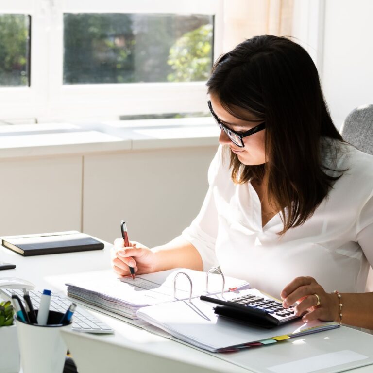 Woman working at a desk with a calculator, writing notes, and organizing documents, emphasizing efficient billing processes for business management.