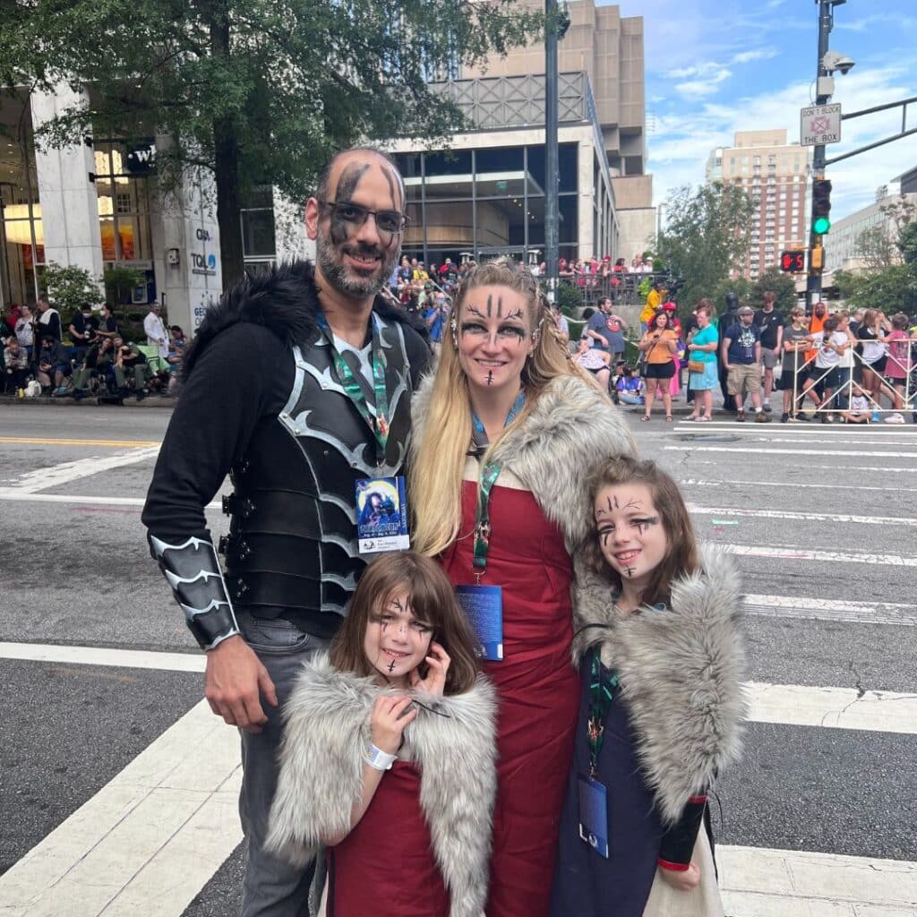 Family dressed in fantasy costumes, featuring a man in a dark, armored outfit, a woman in a red dress with a fur shawl, and two girls in matching outfits, all with face paint, at a lively outdoor event.
