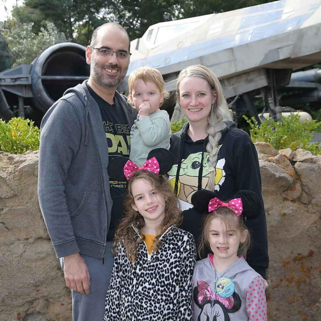 Family portrait featuring Coach Anna Ghanouni with her husband and three children, all smiling and dressed in playful attire, set against a backdrop of a themed attraction, highlighting their enjoyment of family time and outdoor activities.