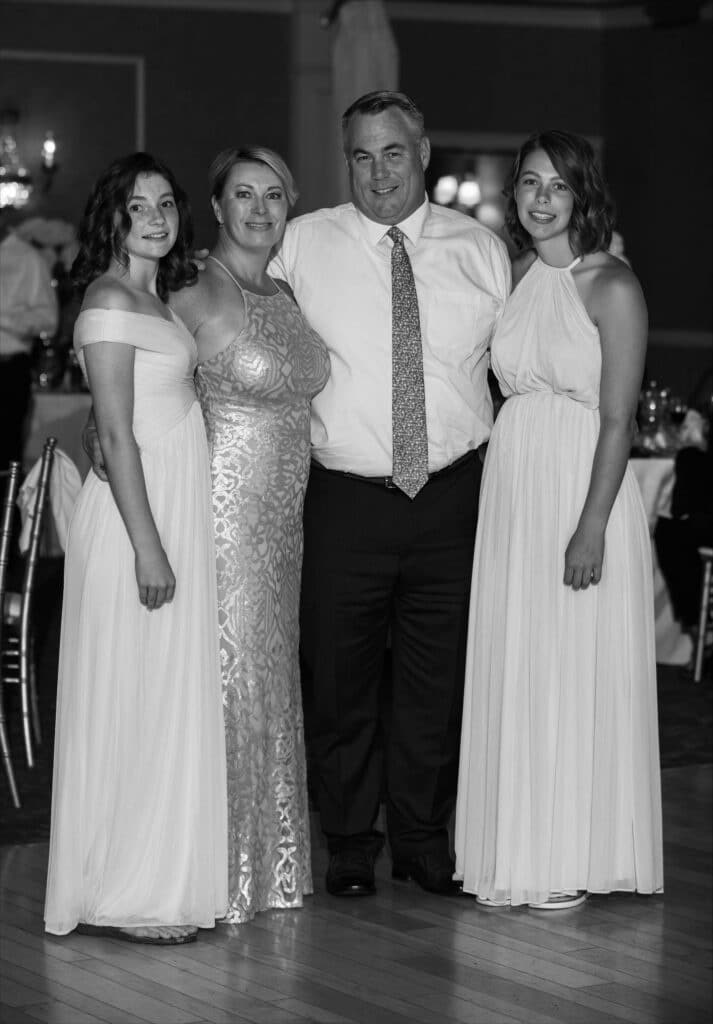 Family portrait of Coach Pamela Coward in a formal setting, wearing a shimmering dress, alongside her husband and two daughters, all dressed in elegant white attire, capturing a moment of togetherness and celebration.