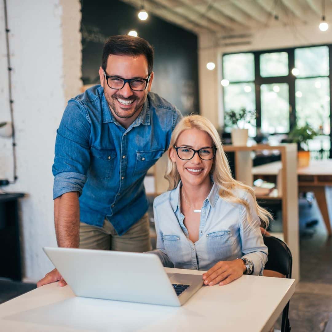 Smiling man and woman in casual attire, collaborating at a laptop in a modern office setting, representing teamwork and support for business owners considering hiring an Executive Assistant.