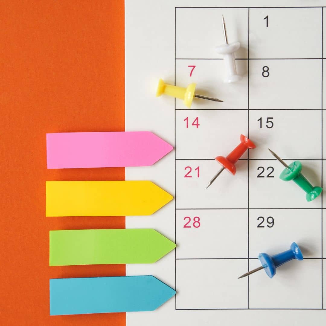 A calendar flatlay with pushpins and sticky tabs on a vivid orange background.