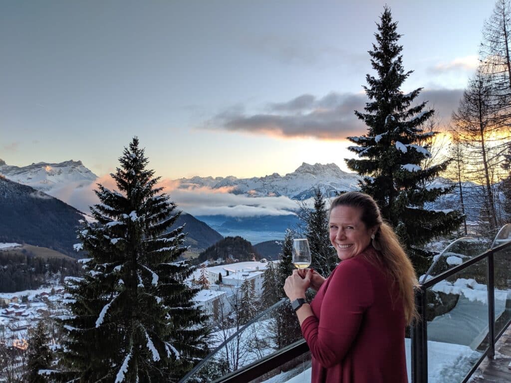 Woman smiling with a glass of wine in front of snow-capped mountains and a sunset, representing a balanced life and success in business coaching.