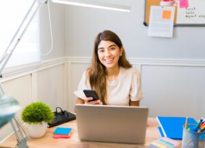 Smiling woman holding smartphone at desk with laptop, emphasizing client engagement and happiness in business strategies.