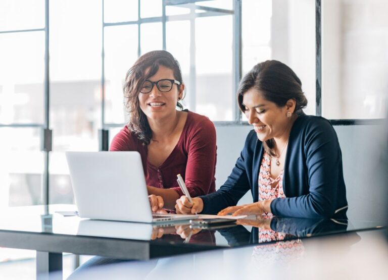 Two women collaborating at a table with a laptop, discussing training strategies for executive assistants to enhance productivity and teamwork.