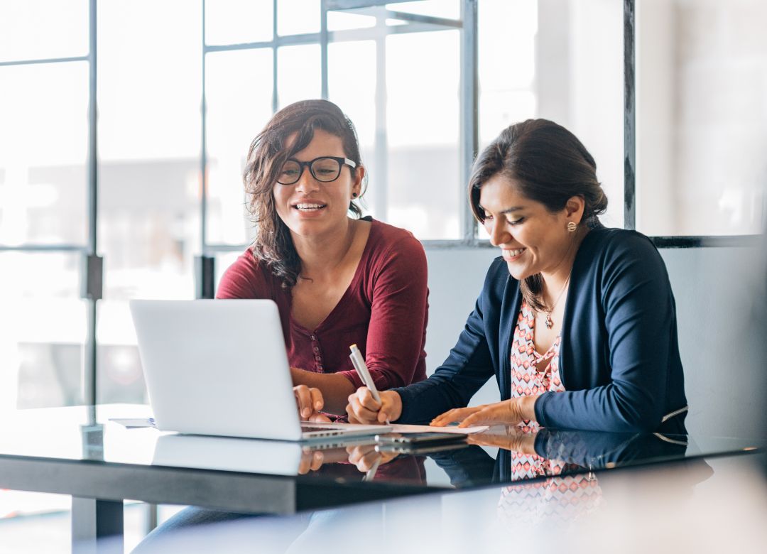 Two women collaborating at a table, one using a laptop and the other taking notes, representing effective training and teamwork for executive assistants.