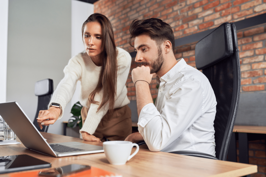 Woman pointing at laptop screen while training a man in a business setting, emphasizing effective communication and collaboration for executive assistant training.