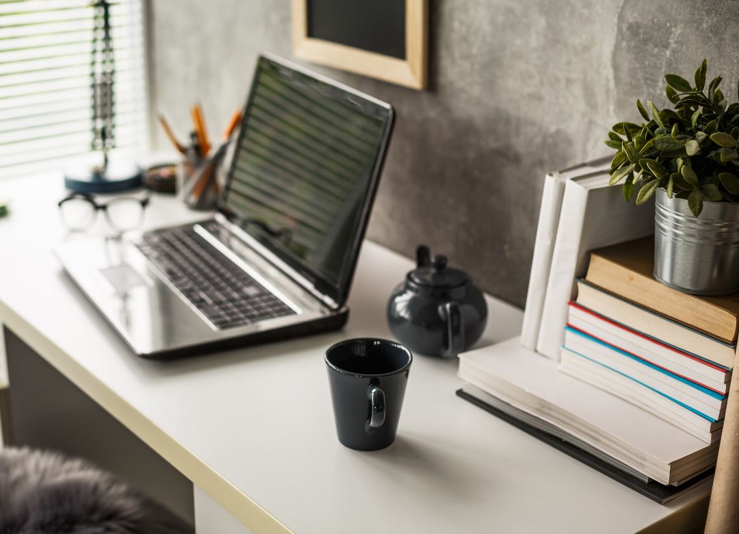 Modern workspace featuring a laptop, a black coffee cup, a teapot, books, and a small potted plant, emphasizing organized business systems and productivity.