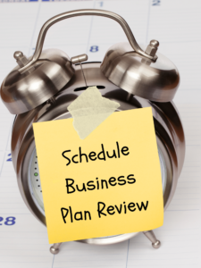 Alarm clock with sticky note saying "Schedule Business Plan Review," emphasizing time management for business planning.