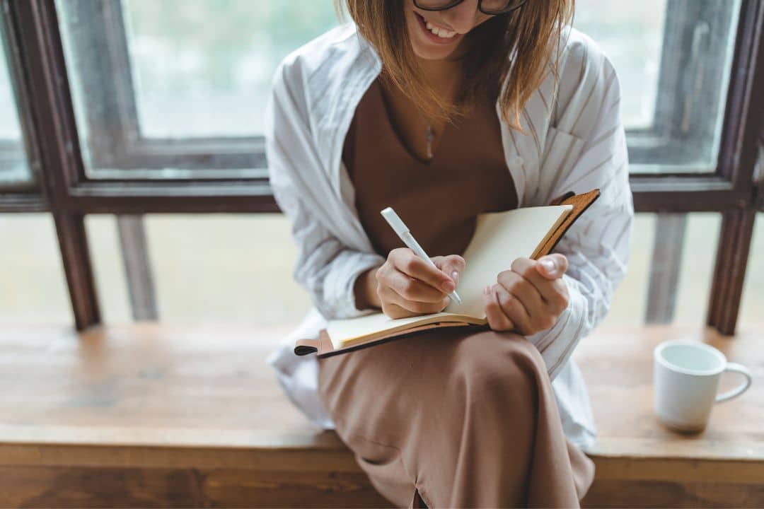 Woman writing in a notebook while sitting by a window, illustrating self-reflection and data-driven leadership in business management.