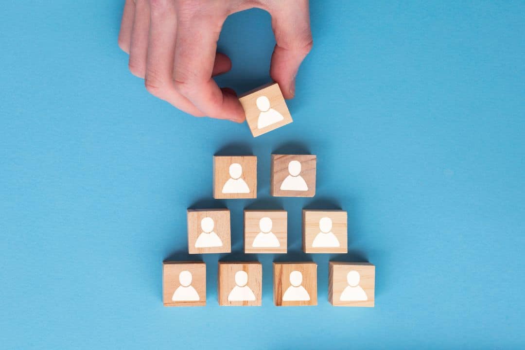 Hand placing wooden block with person icon on top of pyramid structure, symbolizing team building and delegation in business management.