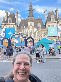 Woman smiling in front of Paris 2024 Olympics signage at a festive outdoor event near the Hôtel de Ville, with people enjoying the atmosphere and vibrant decorations.