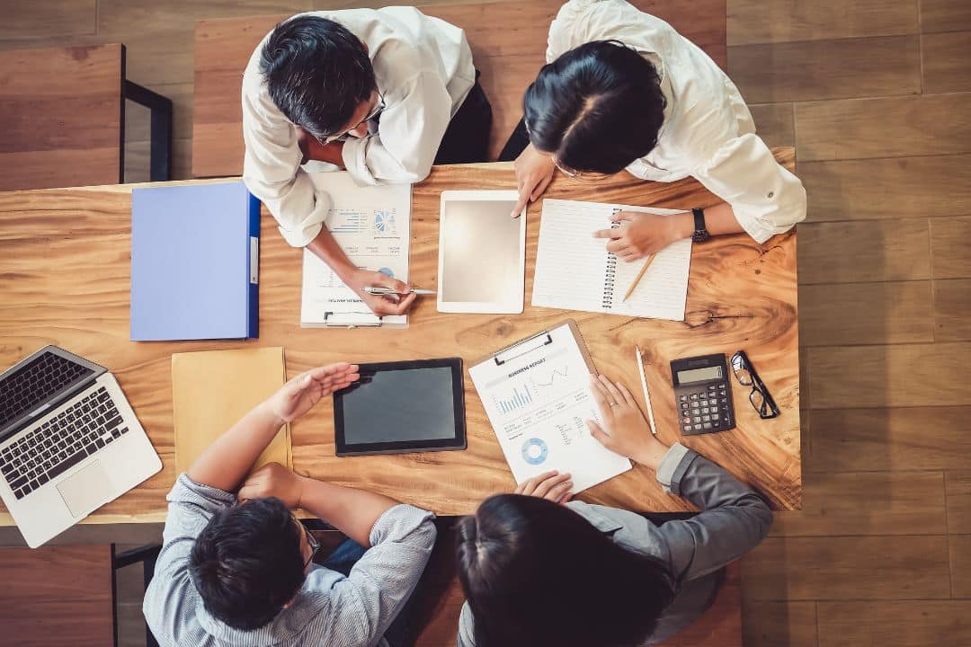 Group of business professionals collaborating at a wooden table with laptops, tablets, documents, and a calculator, emphasizing teamwork and effective business workflows.