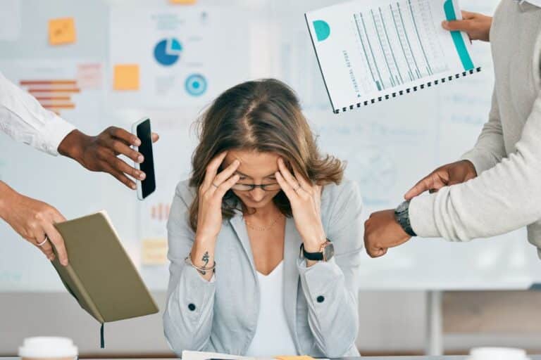 Woman looking stressed at a desk, surrounded by hands holding a smartphone, notebook, and documents, illustrating the challenges of managing multiple tasks in a business environment.