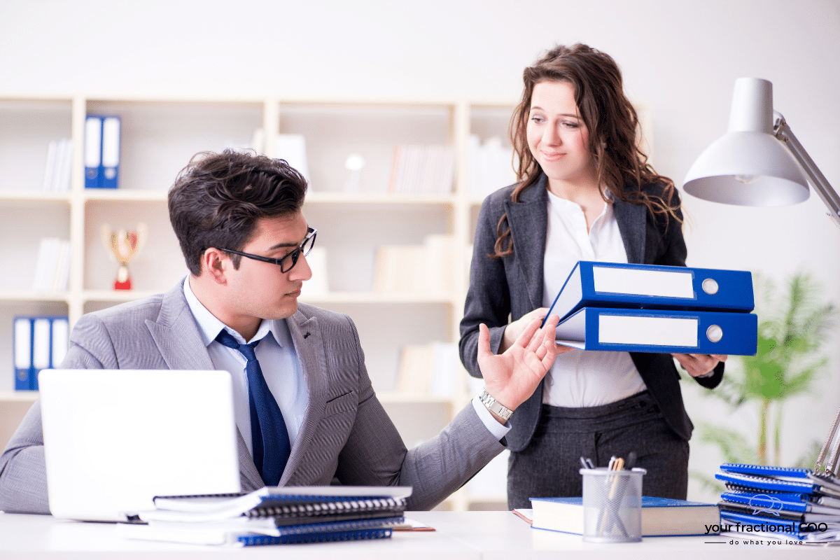Business professionals in an office setting, with a woman in a suit handing over blue folders to a man in formal attire, emphasizing collaboration and task delegation.